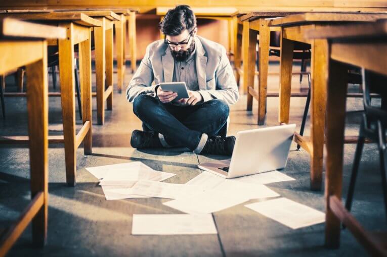 Schülerfrage: Ist das Studium schwer? Student sitzt mit Tablet und Laptop auf dem Boden des Hörsaals, um ihn herum sind Blätter verstreut