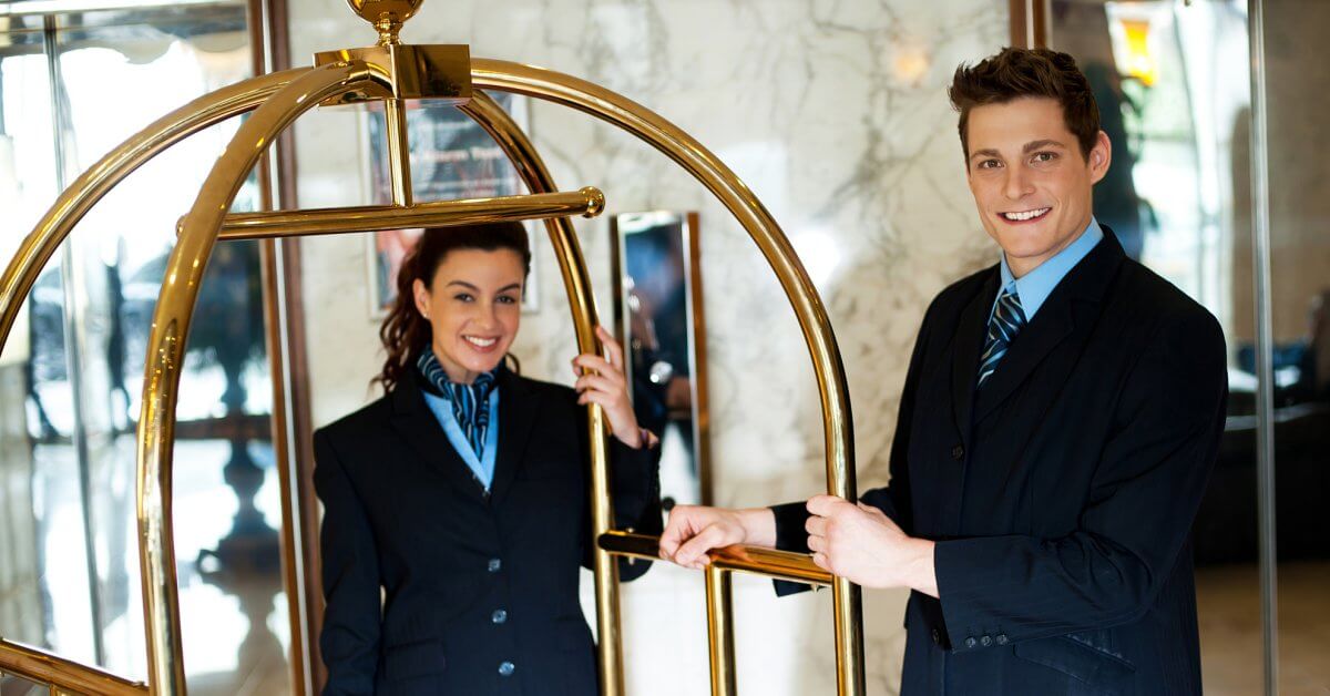 Zwei Junge Hotelmitarbeiter stehen an Kofferwagen und lächeln in Kamera